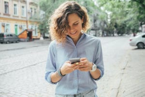 Happy young woman wearing blue shirt using mobile phone and smile on street near road in summer day.