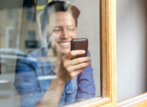Portrait looking through window at smiling young woman using cell phone