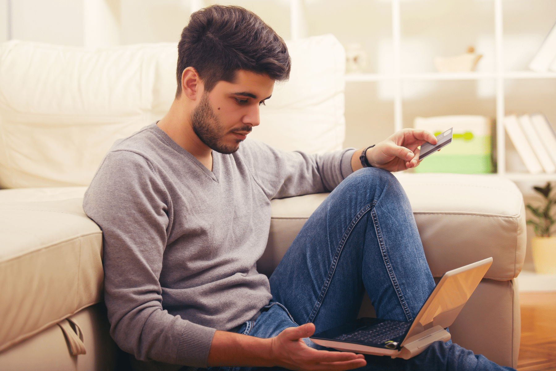 Man sitting at the front of couch looking at computer and holding credit card