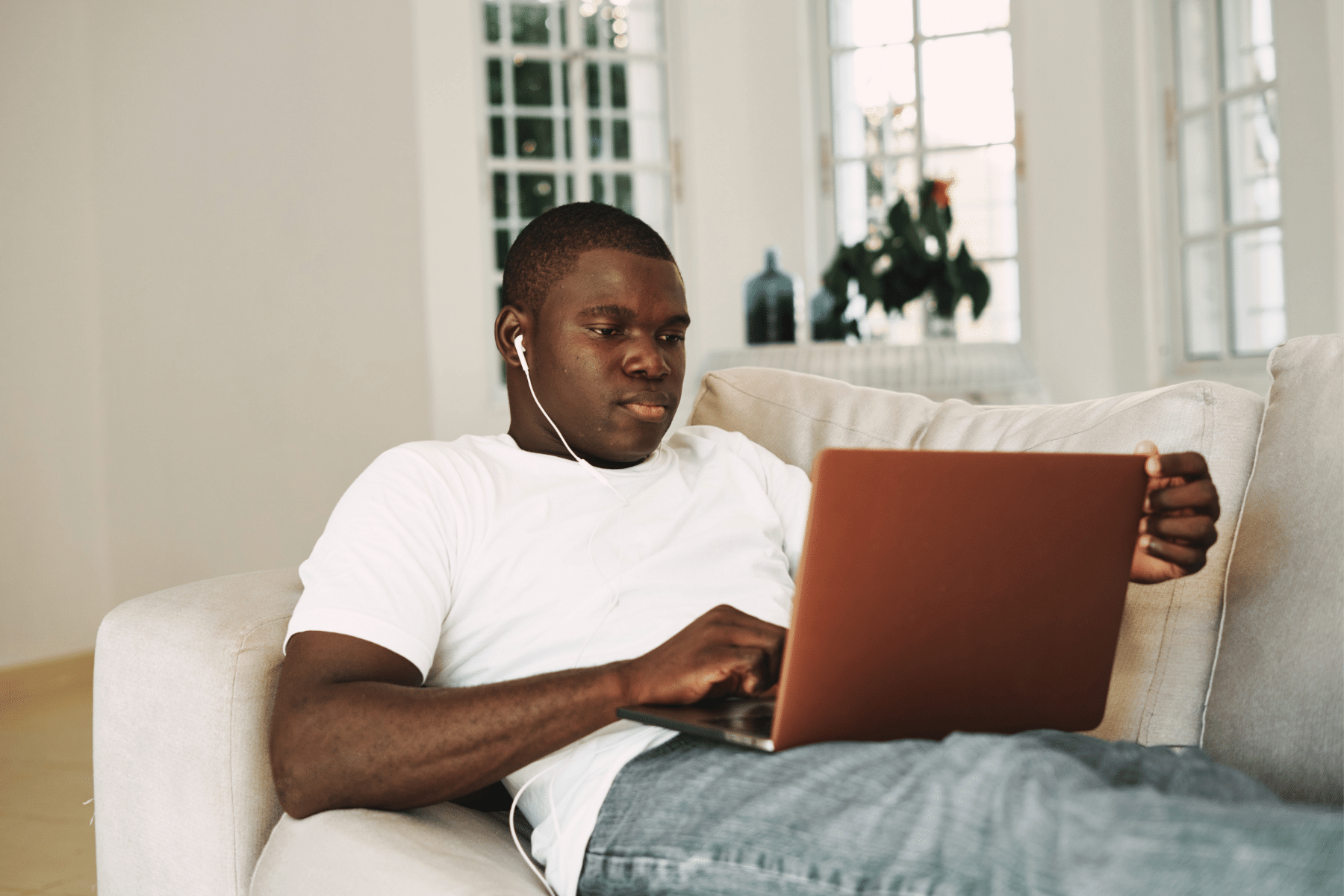 Young man sitting on couch with laptop in his lap while listening through headphones