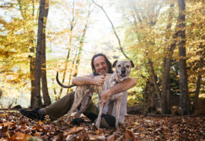 An adult man walks with a dog in the forest. They sit on the grass and hug.