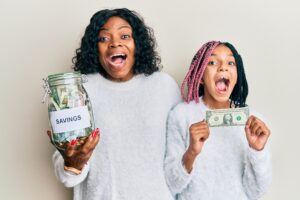Beautiful african american mother and daughter holding jar with savings and 1 dollar banknote celebrating crazy and amazed for success with open eyes screaming excited.
