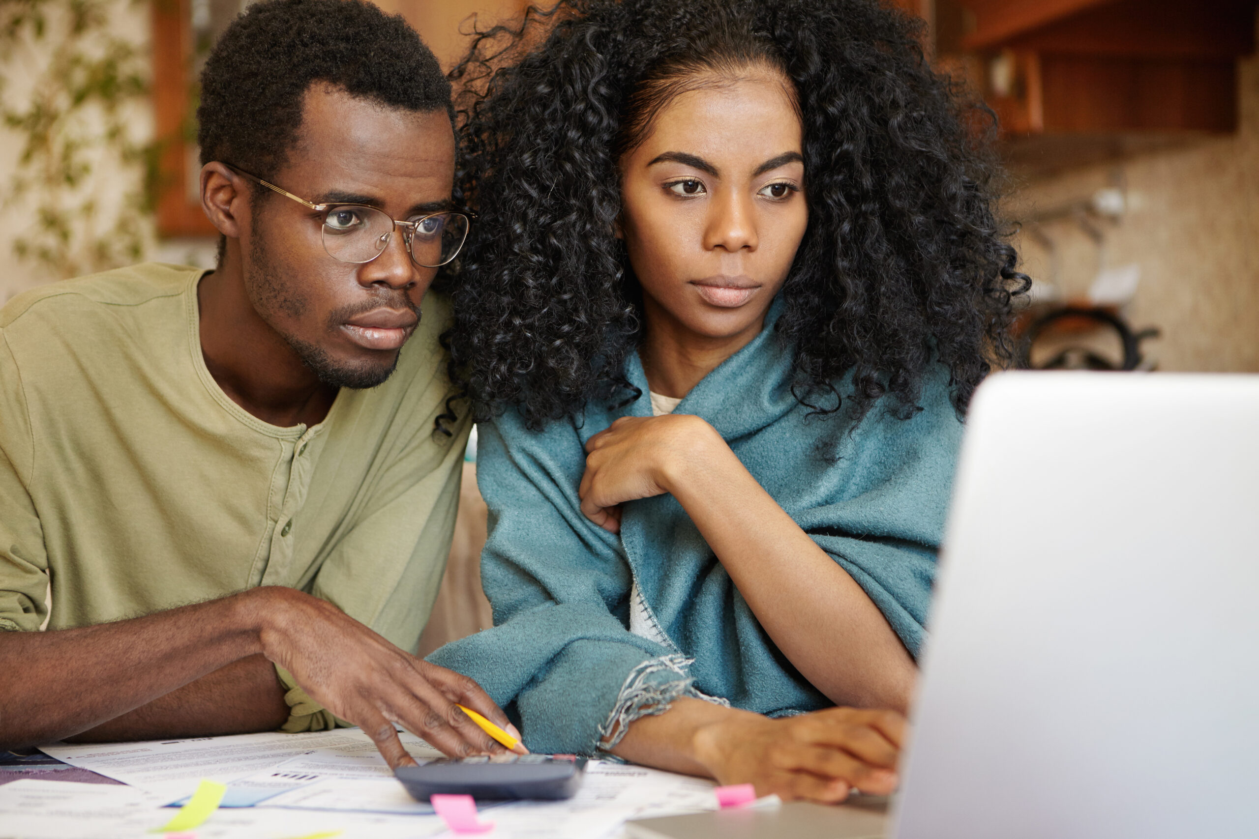 Close-up candid shot of young dark-skinned couple sitting in front of open laptop computer while paying gas and electricity bills online, using banking application, having concentrated looks