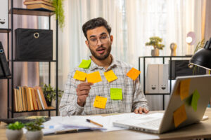 Tired exhausted fatigue young Indian businessman office employee manager sits in home office table with laptop sticky notes with many tasks inscription help, feels sad boredom overworked multitasking.